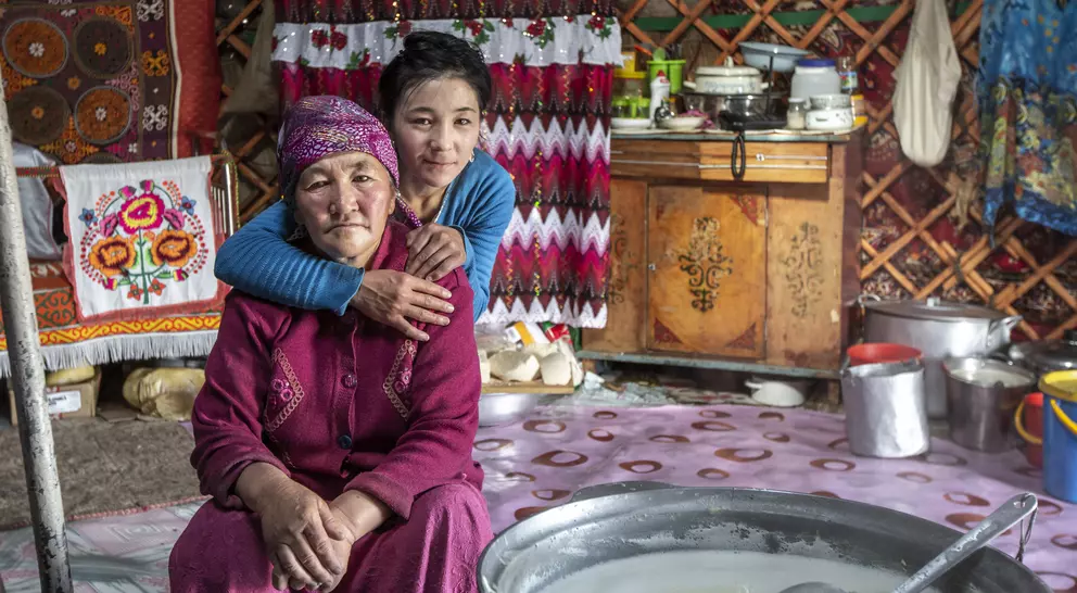 A young girl embraces an elderly woman in a cozy, traditional yurt adorned with colorful textiles and kitchen items.