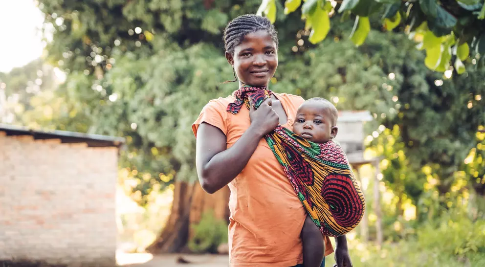 A woman wearing an orange top holds a baby wrapped in colorful fabric, standing outdoors near a home with trees in the background.