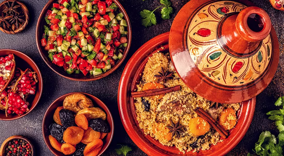 A colorful spread of couscous with spices, surrounded by salads, dried fruits, and garnishes on a textured surface.