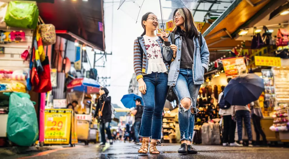 Two friends walk together under an umbrella in a vibrant market, surrounded by colorful stalls and wet pavement.