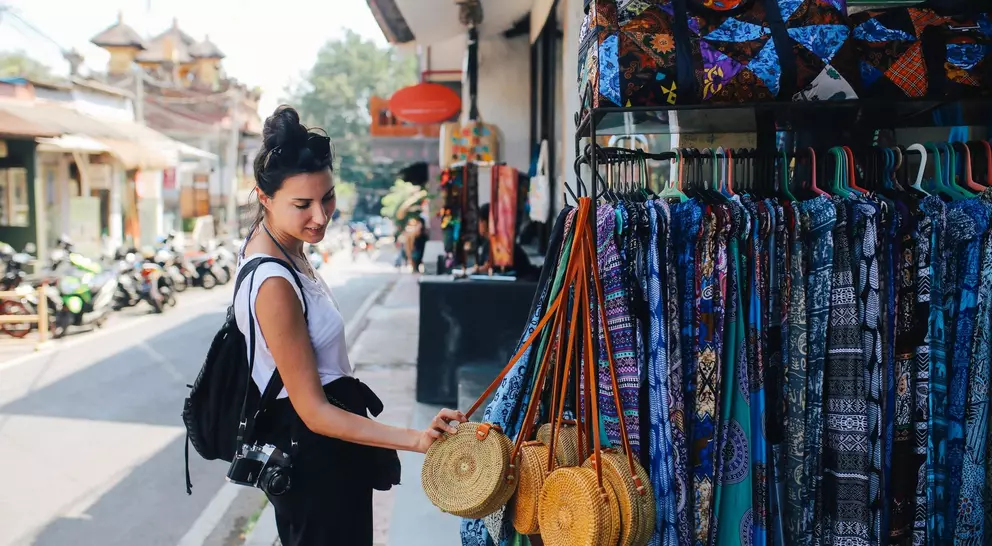 A woman examines woven bags in a market, with colorful fabrics hanging nearby and a street scene in the background.