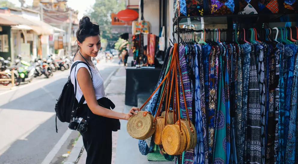 A woman examines woven bags in a market, with colorful fabrics hanging nearby and a street scene in the background.