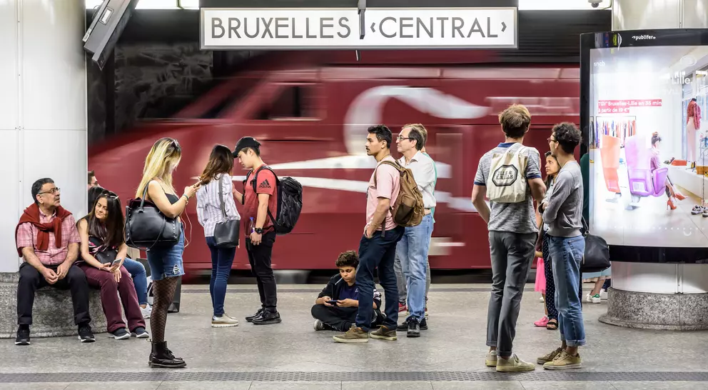 A busy train station platform at Bruxelles Central, with passengers waiting and a train passing in the background.