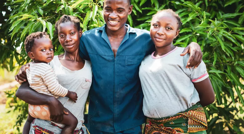 A smiling man stands with three women, one holding a baby, in front of lush green foliage.