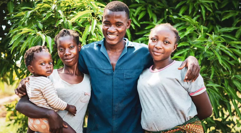 A smiling man stands with three women, one holding a baby, in front of lush green foliage.