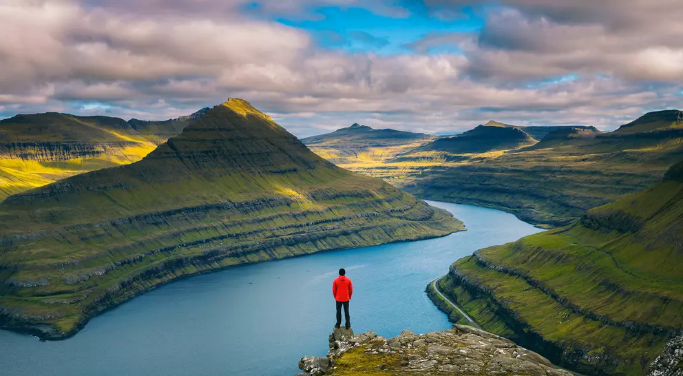 A person in a red jacket stands on a cliff overlooking a winding river and lush green mountains under a cloudy sky.