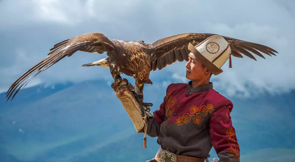 A man in traditional attire holds a large eagle on his arm against a mountainous backdrop.