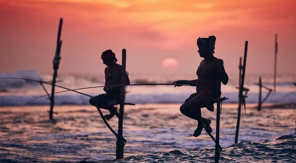Silhouetted fishermen perched on poles in the ocean at sunset, casting lines into the water.
