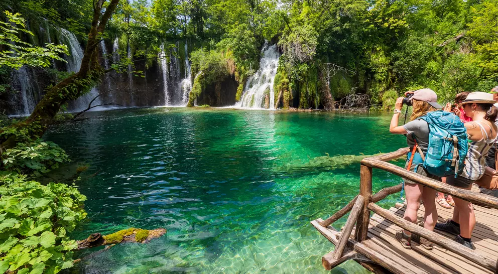 Two people stand on a wooden platform, taking photos of a turquoise lake and waterfalls surrounded by lush greenery.