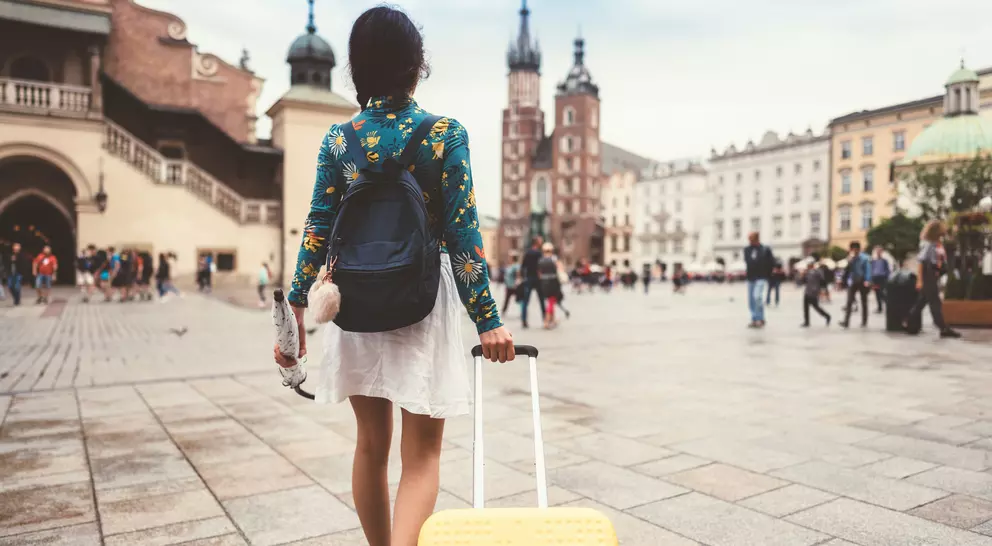 A person walks away, pulling a yellow suitcase through a bustling square with historic buildings in the background.