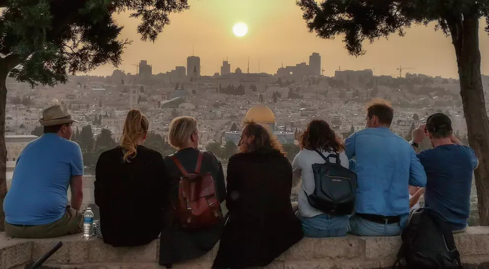 A group of people sit on a stone wall, watching the sunset over a city skyline. Trees frame the scene.