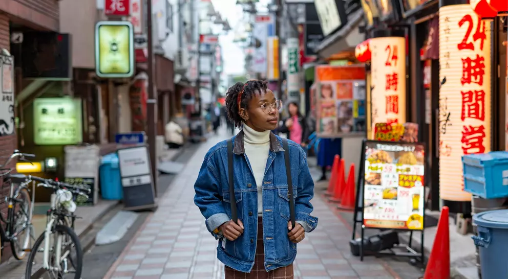 A person in a denim jacket stands in a vibrant urban alley filled with shops and neon signs.