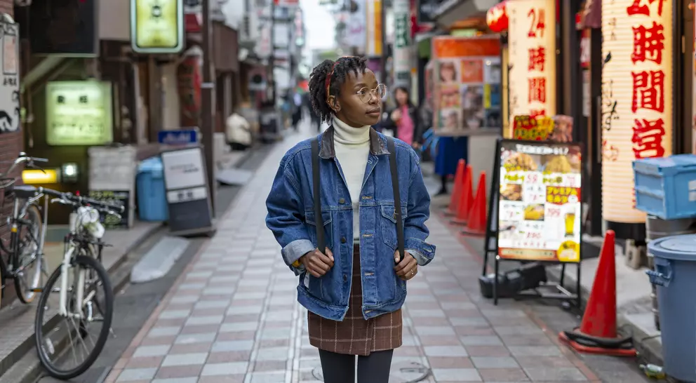 A person in a denim jacket stands in a vibrant urban alley filled with shops and neon signs.