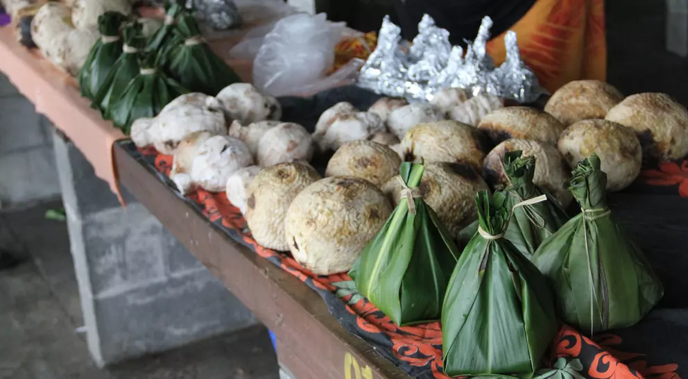 A market stall displays various food items, including round dumplings and bundled parcels wrapped in green leaves.