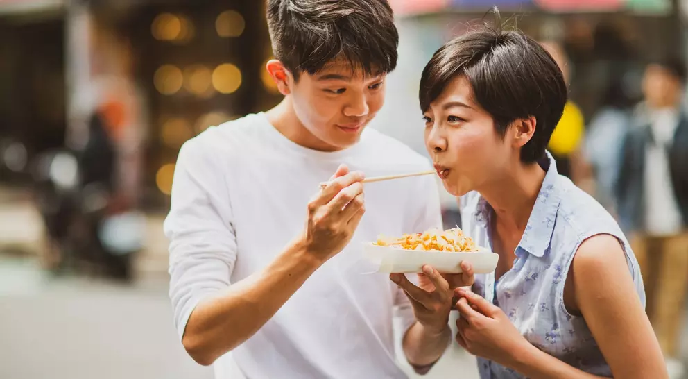 A young man feeding a young woman noodles on a bustling street, both smiling and enjoying the moment.