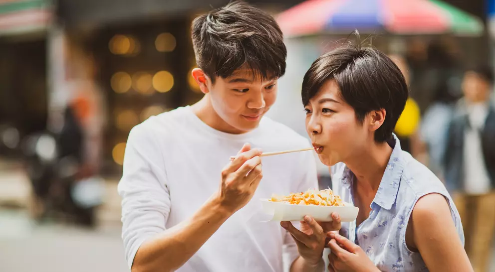 A young man feeding a young woman noodles on a bustling street, both smiling and enjoying the moment.