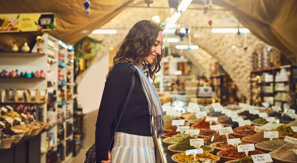 A woman stands in a market, admiring colorful spice displays in bowls, surrounded by shelves filled with various products.
