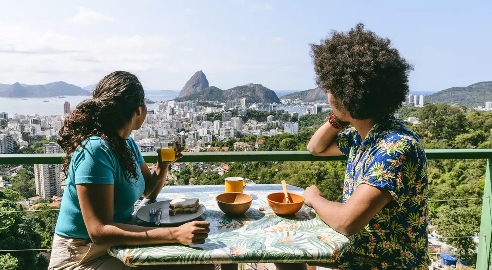 Two people sit at a table with bowls and drinks, overlooking a city and mountainous landscape.
