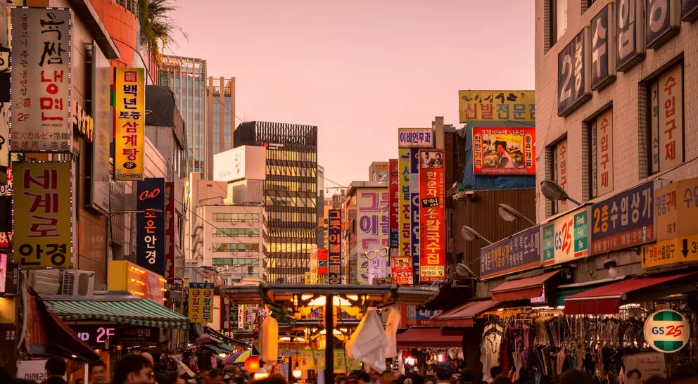 A bustling street market at dusk, filled with colorful signs and people shopping among stalls.