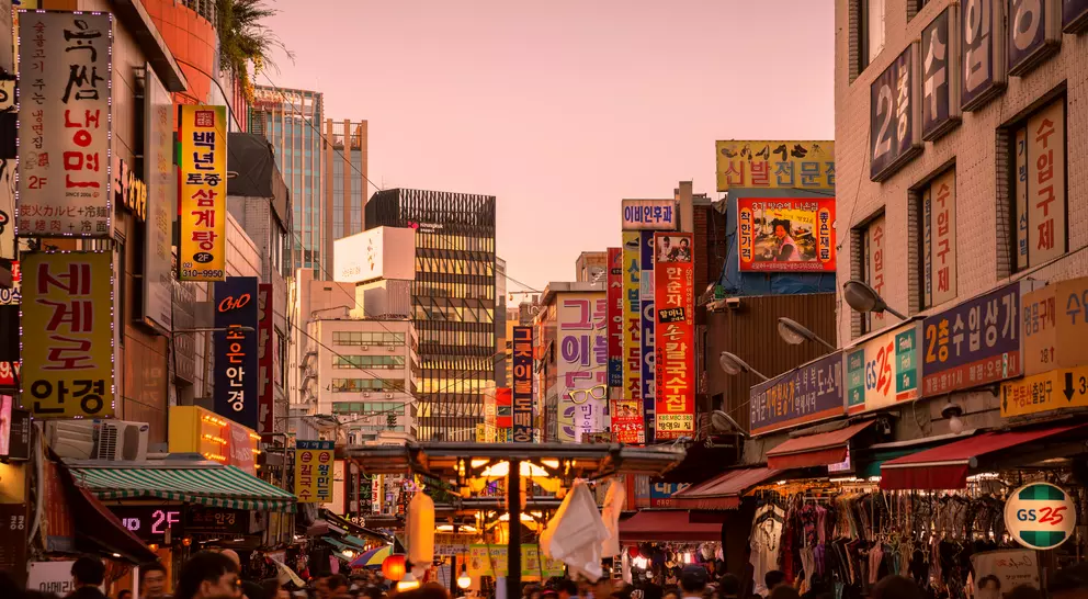 A bustling street market at dusk, filled with colorful signs and people shopping among stalls.