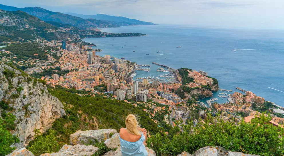 A woman sits on rocks overlooking a coastal city, with buildings and boats nestled along the shoreline and hills in the background.