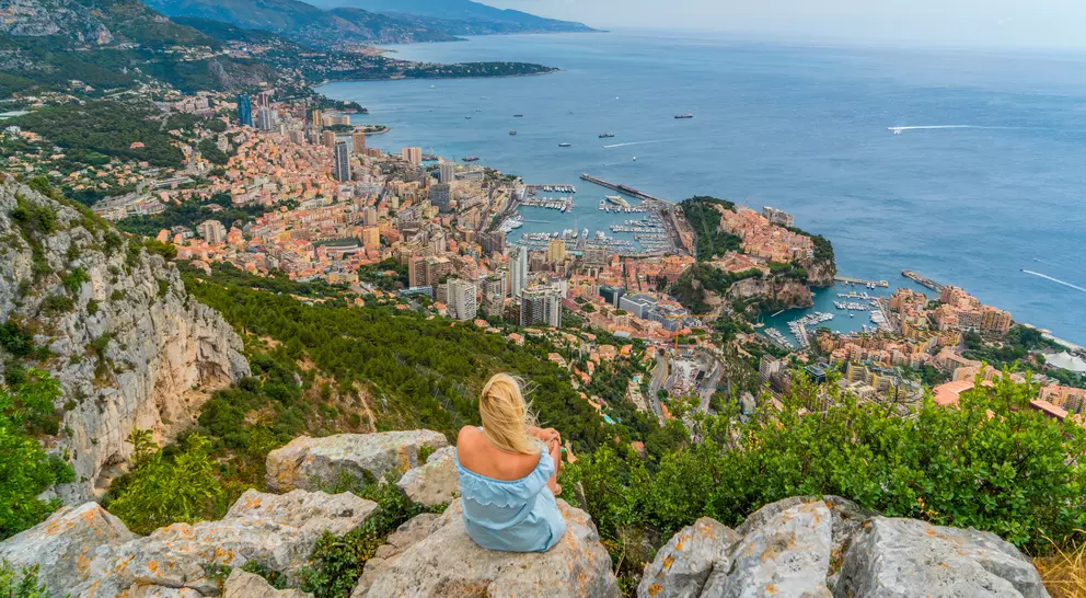 A woman sits on rocks overlooking a coastal city, with buildings and boats nestled along the shoreline and hills in the background.