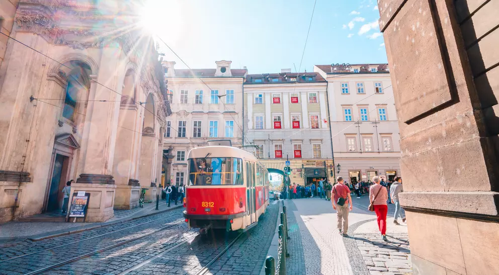 Red tram on a cobblestone street with sun shining, surrounded by historic buildings and people walking.