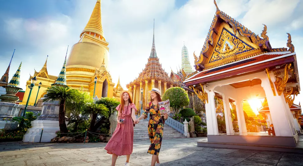 Two smiling women walk hand in hand, surrounded by ornate temples and golden spires, under a bright sky at sunset.