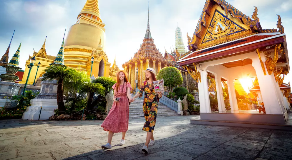 Two smiling women walk hand in hand, surrounded by ornate temples and golden spires, under a bright sky at sunset.
