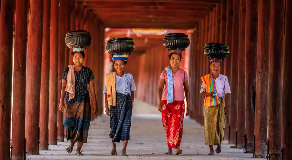 Four girls walk confidently under a wooden archway, each balancing a container on their heads.