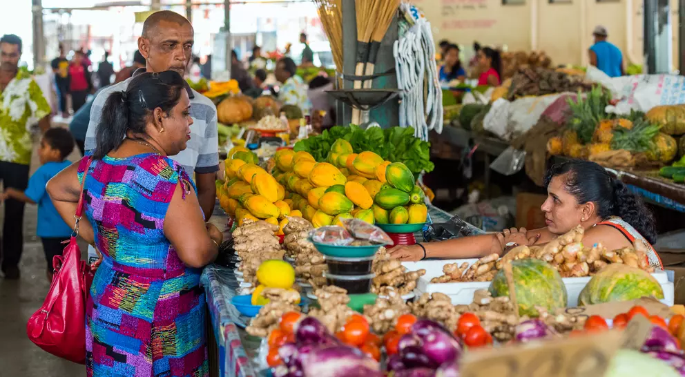 Two women and a man shop at a vibrant market, surrounded by fresh fruits and vegetables in a lively atmosphere.