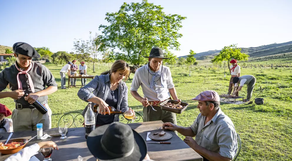 A gathering in a rural setting with people serving food at a table under a clear blue sky and trees.