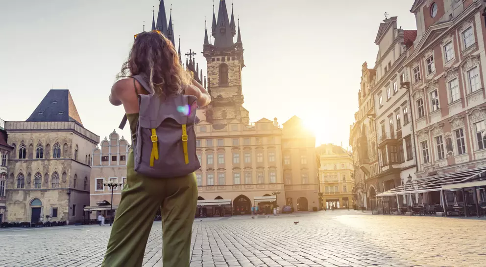 A person with a backpack stands in an empty square, gazing at historic buildings under a golden sunset.