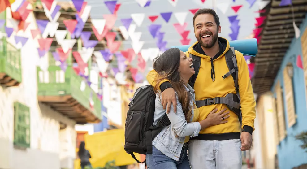 A joyful couple with backpacks laugh together in a colorful street decorated with festive flags.