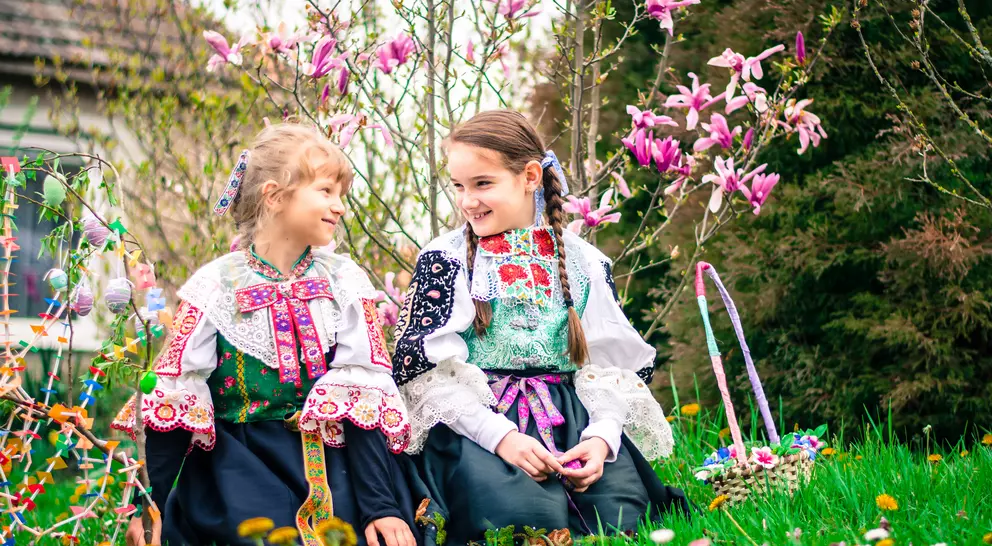 Two girls in traditional costumes sit on the grass, smiling among blooming flowers and plants.