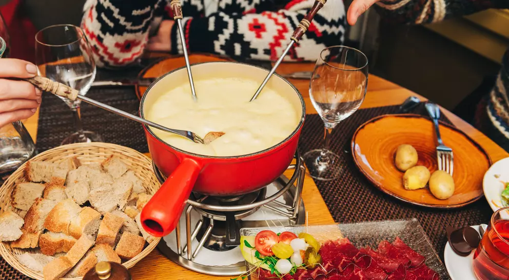 A cheese fondue pot surrounded by bread, vegetables, and drinks, with hands dipping into the fondue.