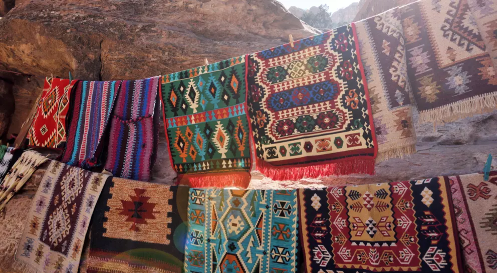 Colorful woven rugs displayed on a rocky surface, showcasing intricate patterns and textures in sunlight.