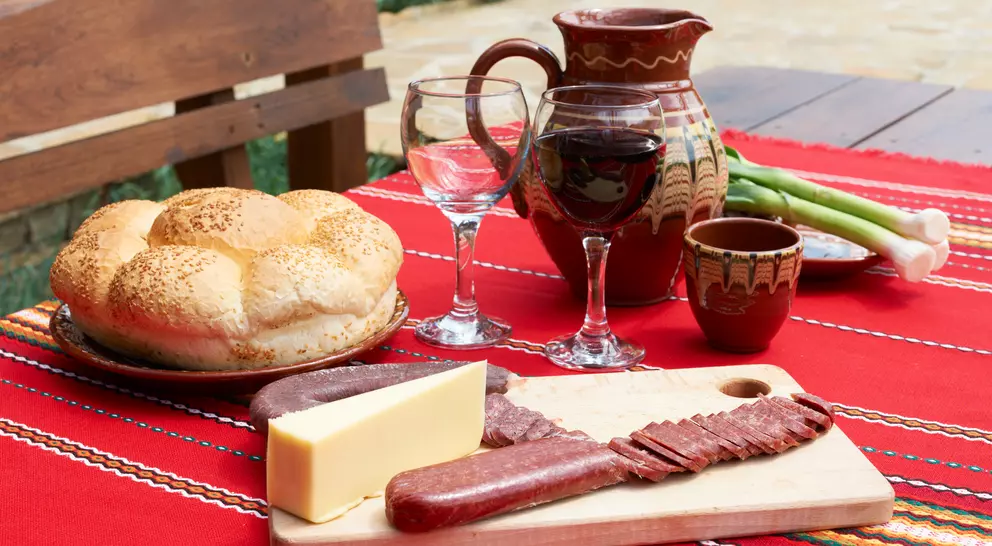 A wooden table set with bread, cheese, cured meats, wine glasses, and a decorated pitcher on a colorful tablecloth.