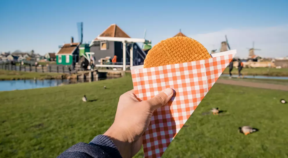 A hand holds a waffle cone with a round pastry, set against a scenic view of a green park and colorful buildings.