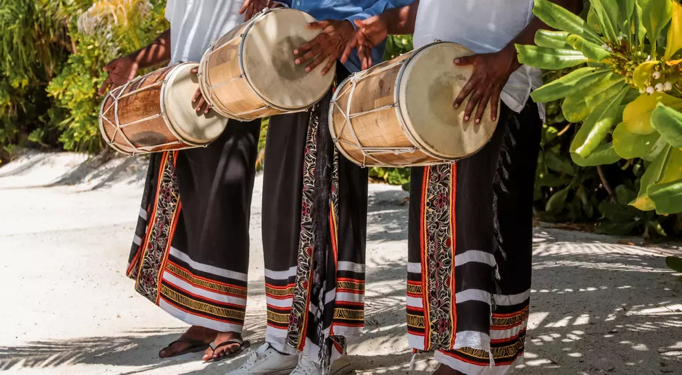 Three musicians in traditional attire playing drums outdoors, surrounded by lush greenery.