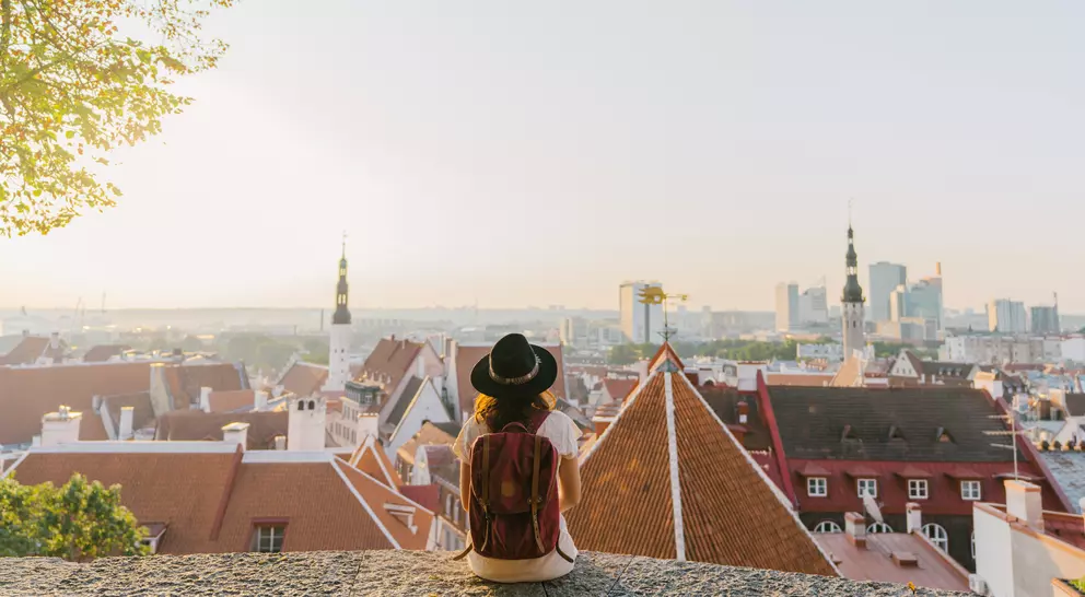 Person in a hat sitting on a ledge, overlooking a cityscape of rooftops and buildings at sunset.
