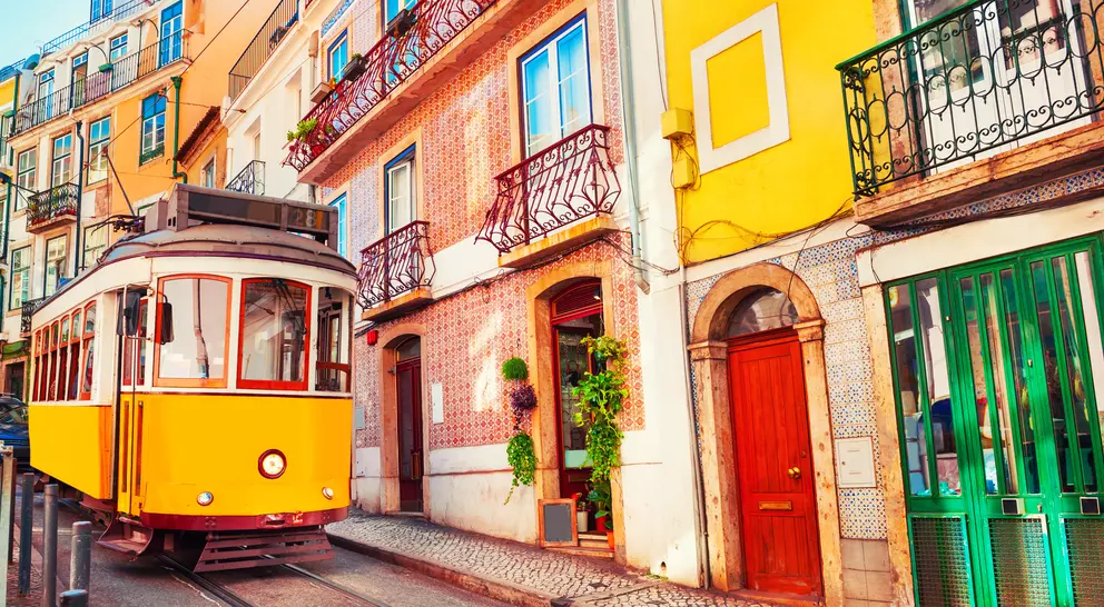 A vintage yellow tram travels along a cobblestone street lined with colorful buildings and balconies in a scenic neighborhood.