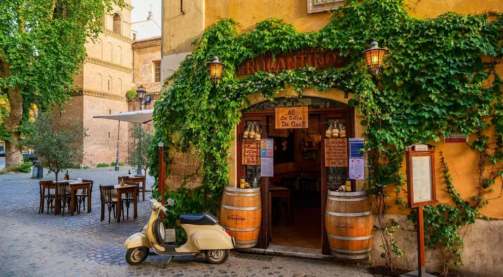 Charming outdoor café entrance with greenery, a vintage scooter, and rustic wooden tables in a cobblestone square.
