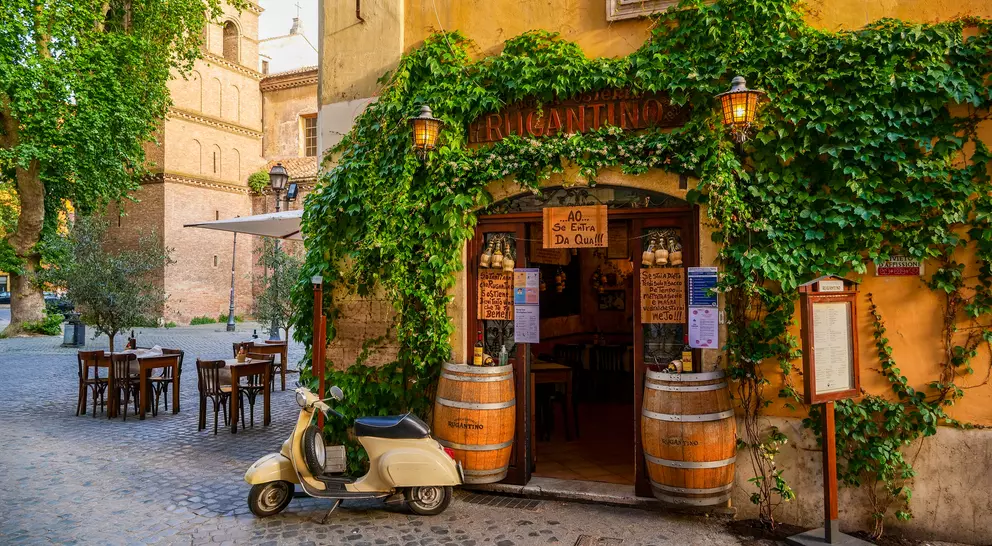Charming outdoor café entrance with greenery, a vintage scooter, and rustic wooden tables in a cobblestone square.