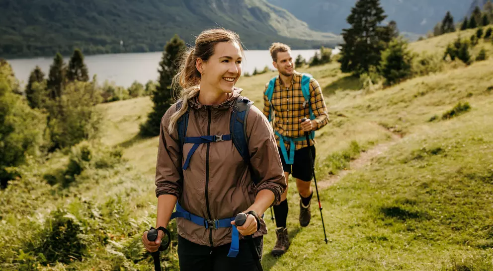 A woman and man hike on a grassy trail with mountains and a lake in the background, enjoying nature.