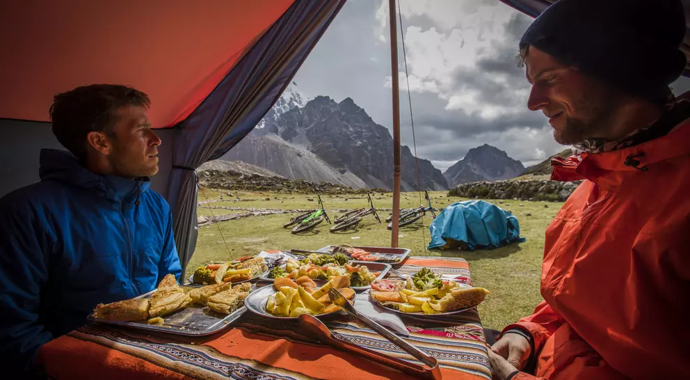 Two people sit inside a tent with a table of food, overlooking a scenic mountain landscape.