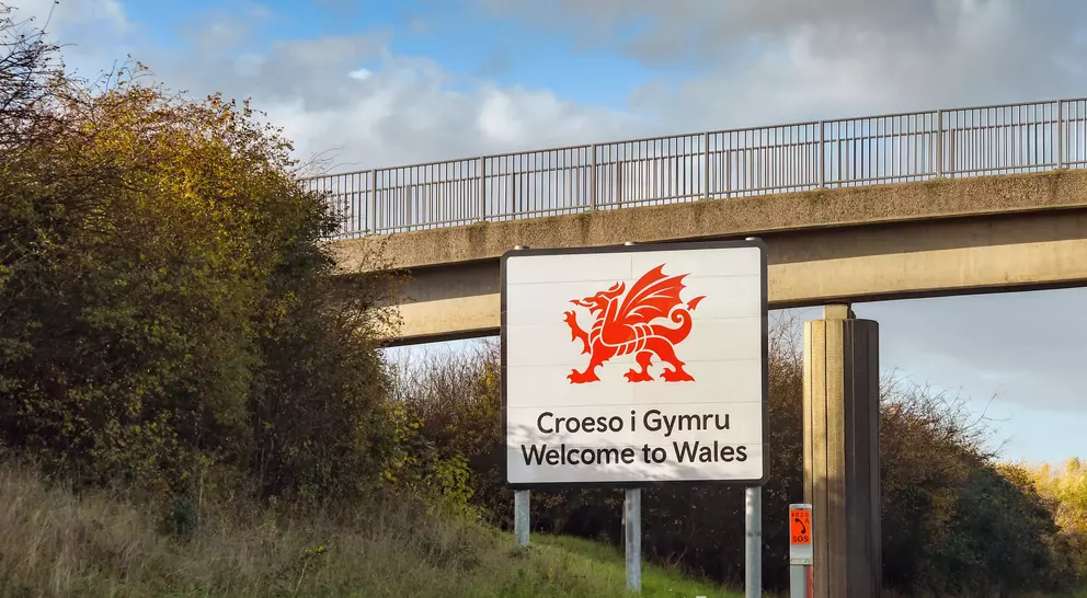 Sign welcoming visitors to Wales with a red dragon emblem, bordered by greenery and a bridge overhead.