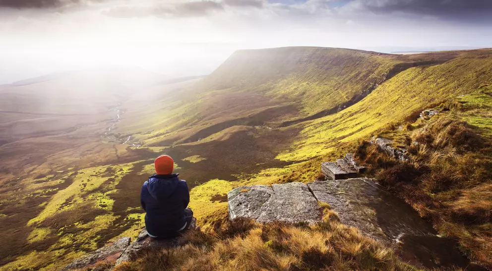 Person sitting on a rocky ledge, gazing at a vast green valley under a cloudy sky.