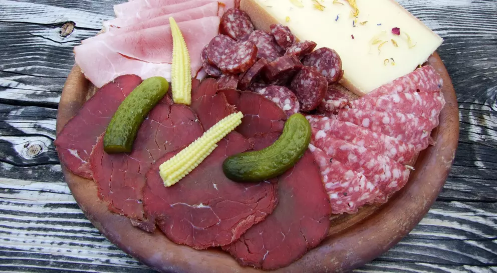 A wooden platter with assorted meats, cheeses, and pickles, including ham, salami, and baby corn.