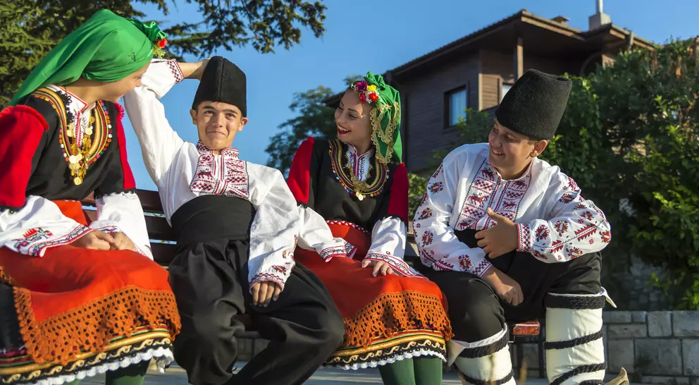 Four young people in traditional Bulgarian clothing pose together outdoors, smiling and engaging with each other.